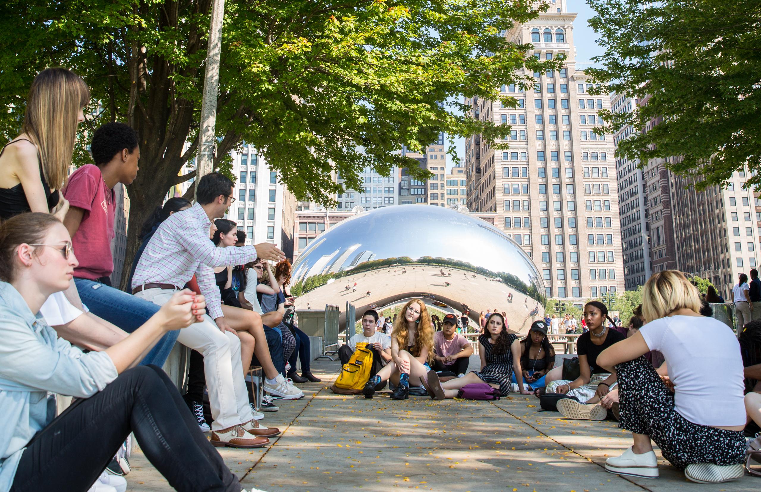 students sitting outside on a sunny day near bean shaped chrome reflective sculpture Cloud Gate in Milennnium Park